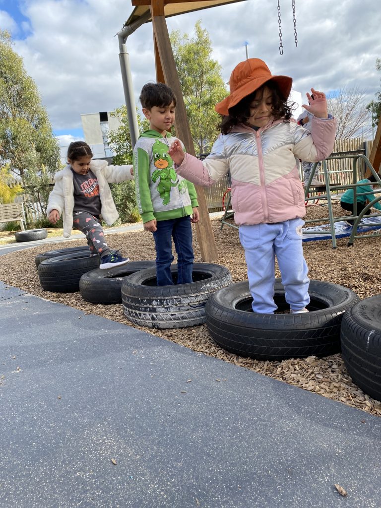 alt: "2 girls and one boy jumping inside car tires obstacle course as a part of risky play."